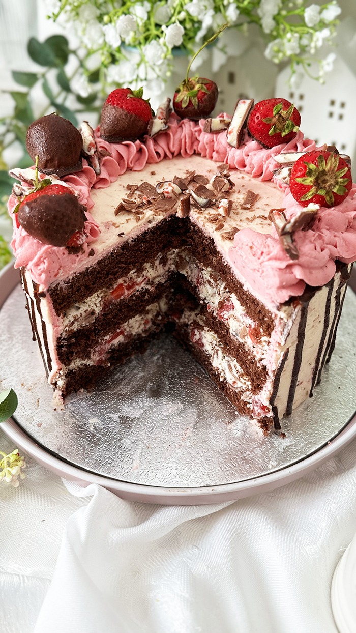 Das Bild zeigt eine zweischichtige Yogurette-Torte, dekoriert mit rosa Sahne, Schokoladen-Drip, in Schokolade getauchten Erdbeeren und Yogurette-Stückchen, umgeben von Blumen