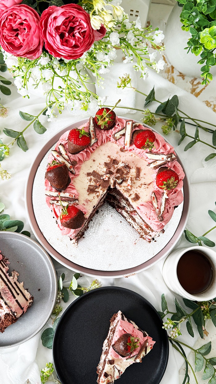 Das Bild zeigt eine zweischichtige Yogurette-Torte, dekoriert mit rosa Sahne, Schokoladen-Drip, in Schokolade getauchten Erdbeeren und Yogurette-Stückchen, umgeben von Blumen
