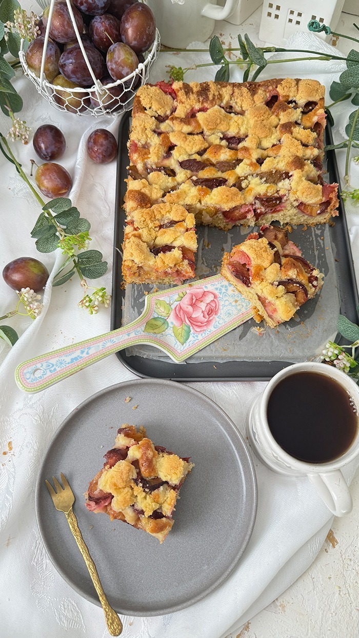 Auf dem Bilde erblicket man einen köstlichen Zwetschgenkuchen mit Streusel mit goldbrauner Streuseldecke und saftigen Früchten. Ein Stück liegt auf einem Teller, während der restliche Kuchen im Hintergrund in der Form ruht, umrahmt von zarter grüner Dekoration. Ein wahrhaft verlockender Anblick!