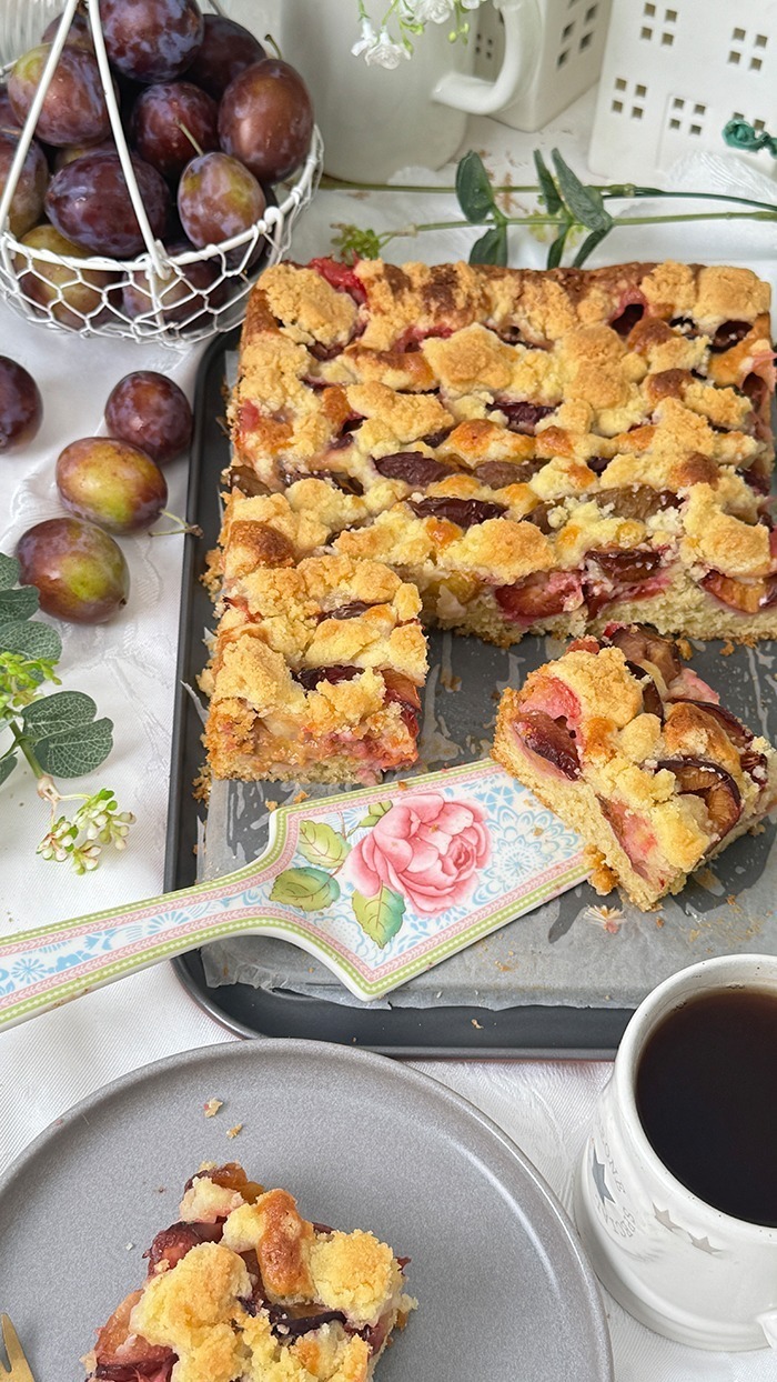 Auf dem Bilde erblicket man einen köstlichen Zwetschgenkuchen mit Streusel mit goldbrauner Streuseldecke und saftigen Früchten. Ein Stück liegt auf einem Teller, während der restliche Kuchen im Hintergrund in der Form ruht, umrahmt von zarter grüner Dekoration. Ein wahrhaft verlockender Anblick!