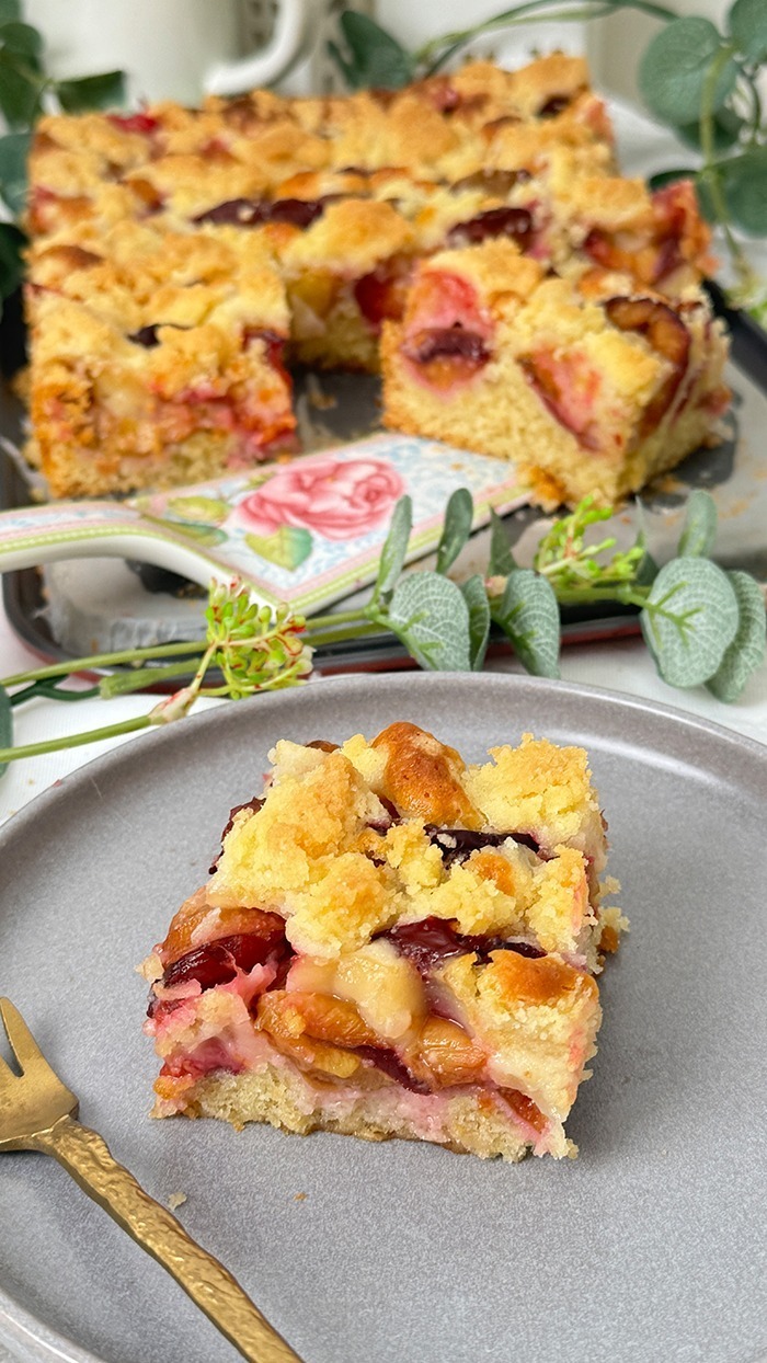 Auf dem Bilde erblicket man einen köstlichen Zwetschgen-Streuselkuchen mit goldbrauner Streuseldecke und saftigen Früchten. Ein Stück liegt auf einem Teller, während der restliche Kuchen im Hintergrund in der Form ruht, umrahmt von zarter grüner Dekoration. Ein wahrhaft verlockender Anblick!