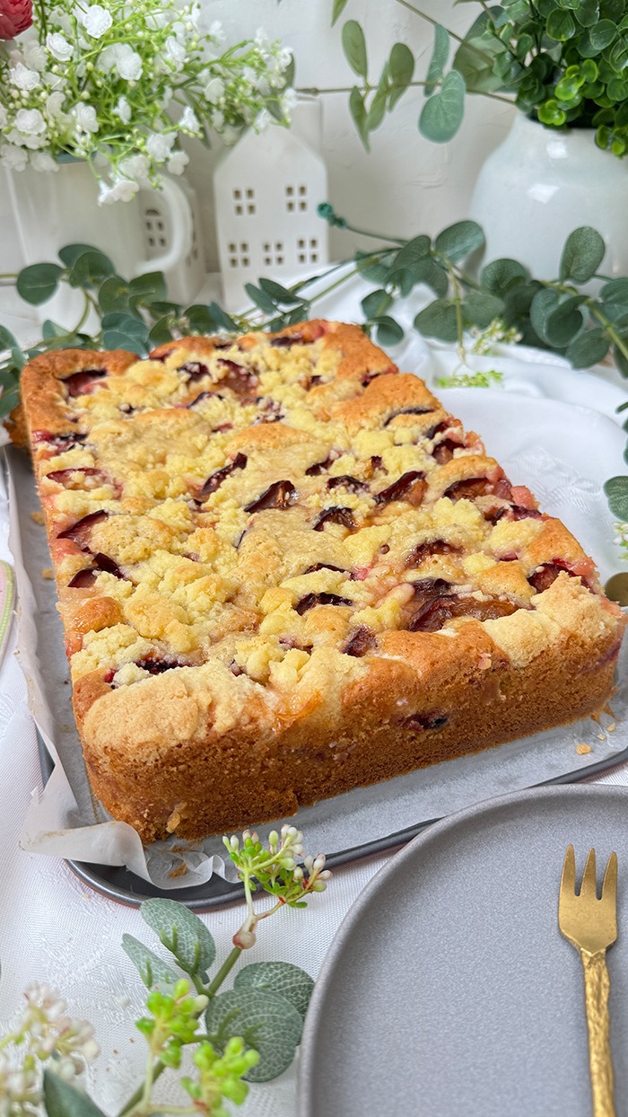 Auf dem Bilde erblicket man einen köstlichen Zwetschgenkuchen mit Streusel mit goldbrauner Streuseldecke und saftigen Früchten. Ein Stück liegt auf einem Teller, während der restliche Kuchen im Hintergrund in der Form ruht, umrahmt von zarter grüner Dekoration. Ein wahrhaft verlockender Anblick!