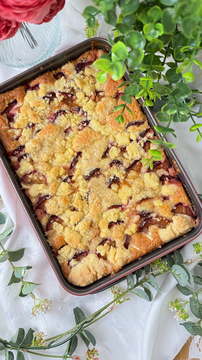 Auf dem Bilde erblicket man einen köstlichen Zwetschgenkuchen mit Streusel mit goldbrauner Streuseldecke und saftigen Früchten. Ein Stück liegt auf einem Teller, während der restliche Kuchen im Hintergrund in der Form ruht, umrahmt von zarter grüner Dekoration. Ein wahrhaft verlockender Anblick!